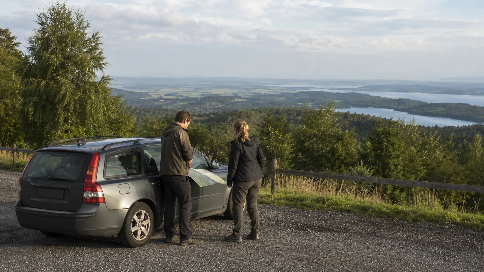 Två resenärer planerar vid en parkerad bil med utsikt över Lygnern och Hallands kustslätt från Fjärås Bräcka