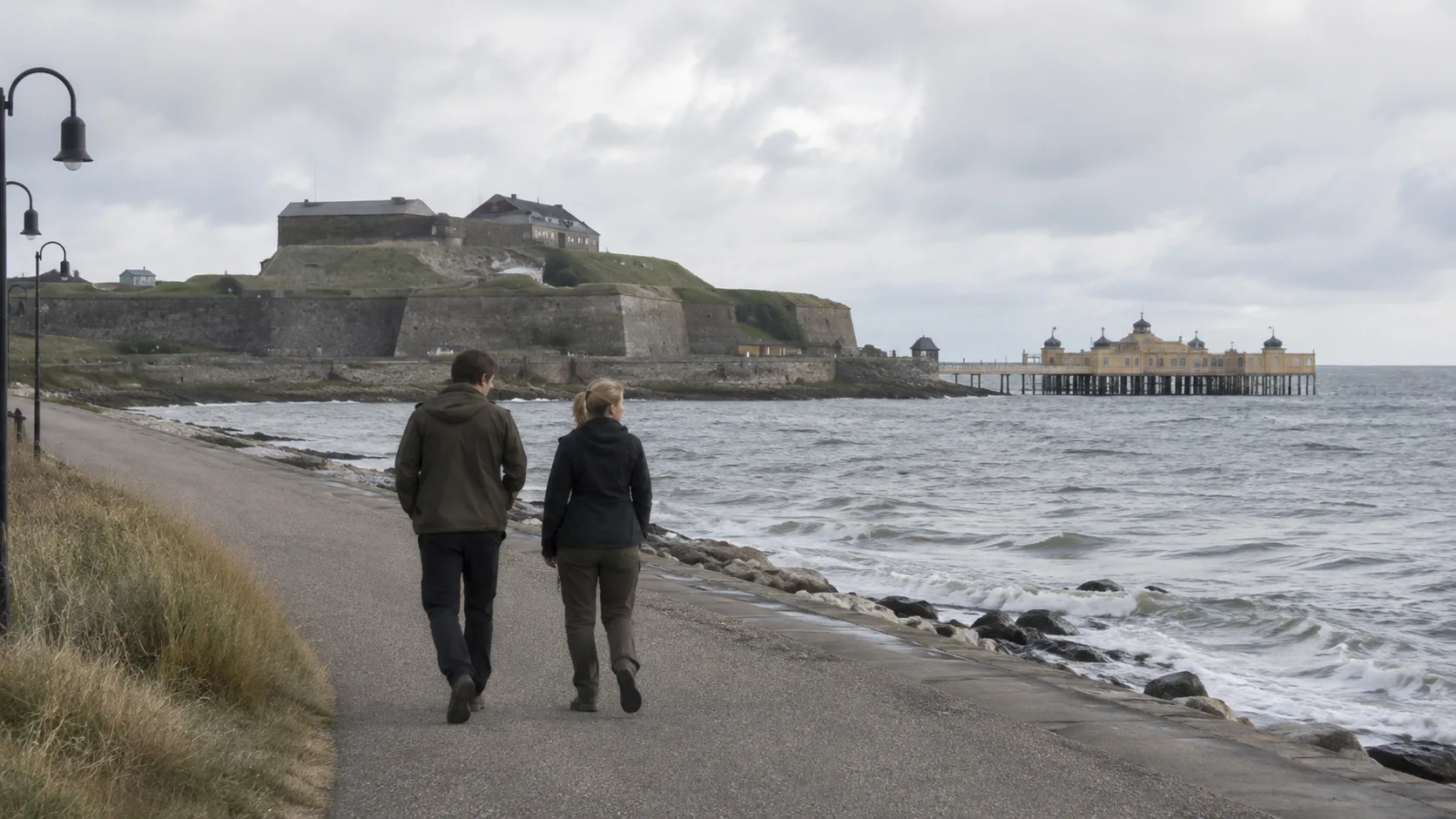 Två resenärer går längs Varbergs strandpromenad med fästningen och Kallbadhuset vid havet