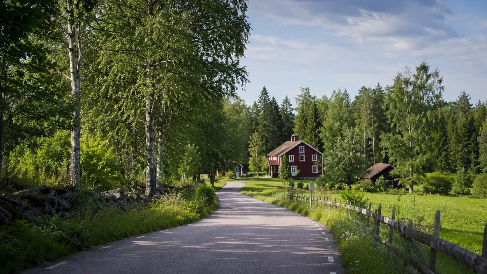 Småväg genom värmländsk skog mot rött hus och kulturmiljö i sommarljus